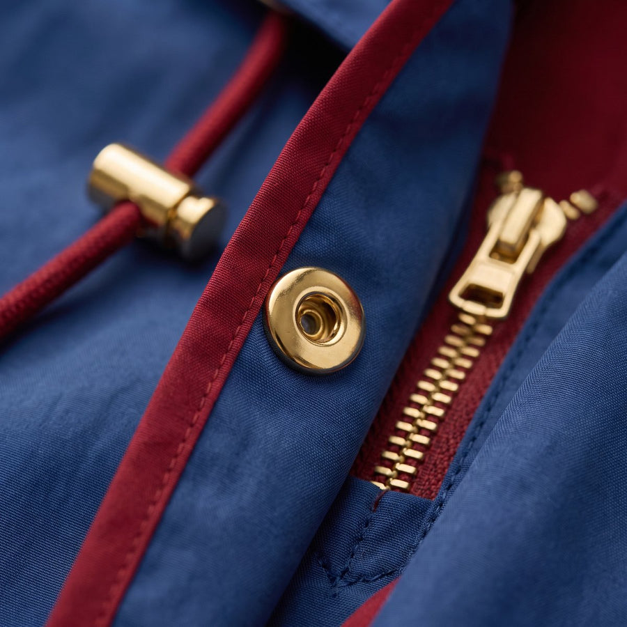 Close-up of gold-tone hardware on blue fabric: a snap, a cord-lock and a front zip