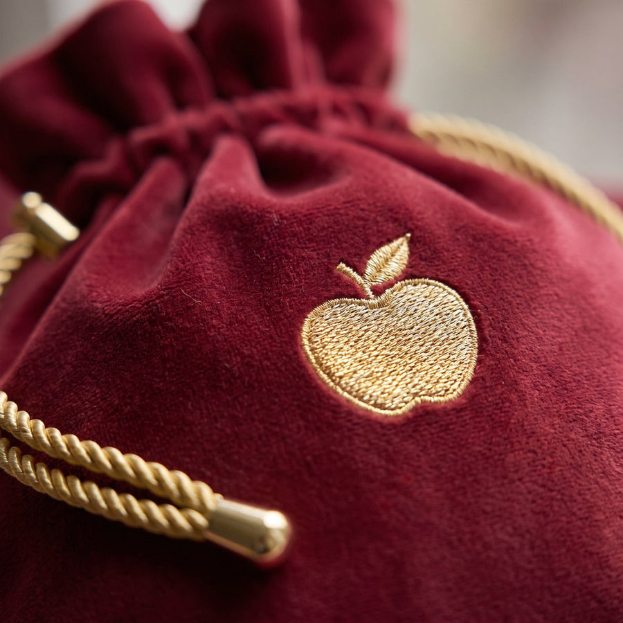 Close-up of a deep-red velour story pouch with a gold embroidered apple and gold drawcord