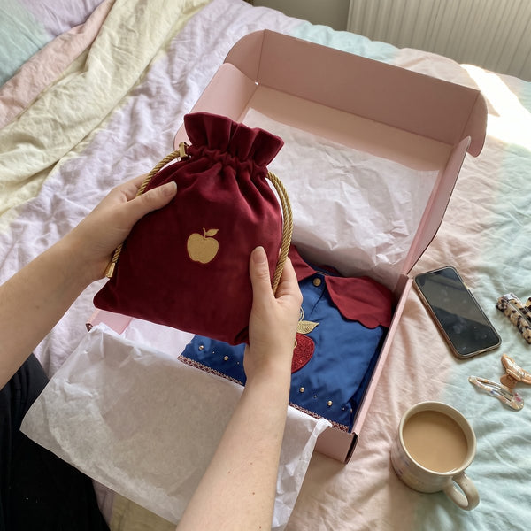 Hands lifting the deep-red FAIREST story pouch out of a pink mailer box on a bed
