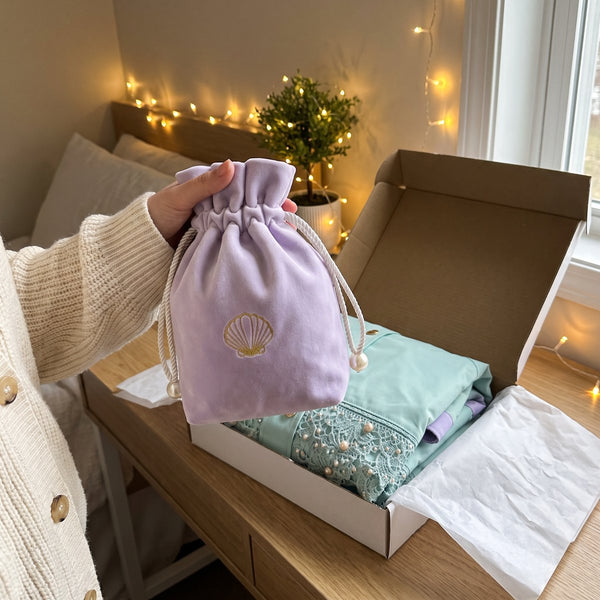 A hand holding up the lilac DEEP SEA story pouch beside an open box with the folded poncho